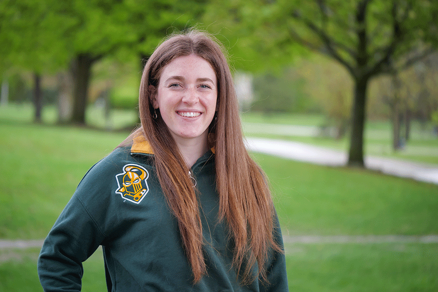 Clarkson Ignite graduate fellow Kelsey Hamilton poses outside on a sunny day in front of trees while wearing a Clarkson athletics logo sweatshirt